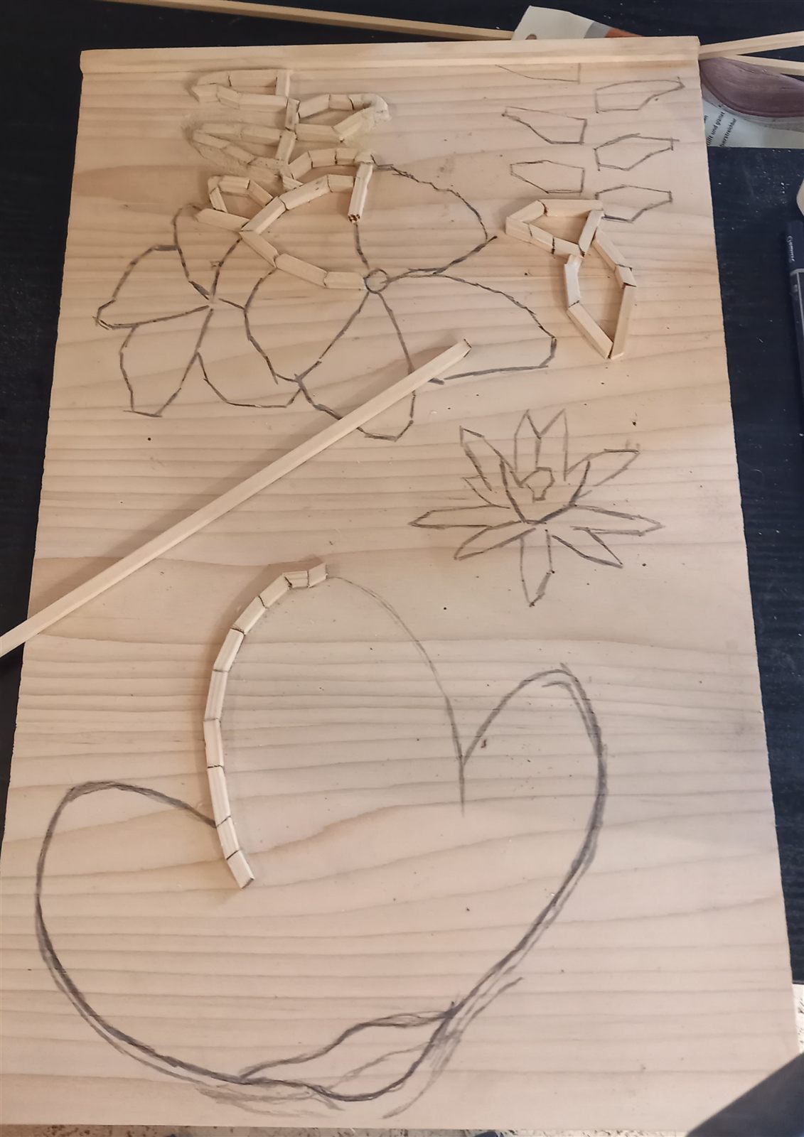 Holzplatte mit vorgezeichneten Blüten und einem Herz, deren Umrisse Stück für Stück aus kleinen Holzstäben gelegt werden